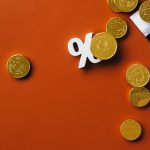 Moedas douradas sobre um gráfico de mercado, ao lado de um símbolo de porcentagem, em fundo laranja.