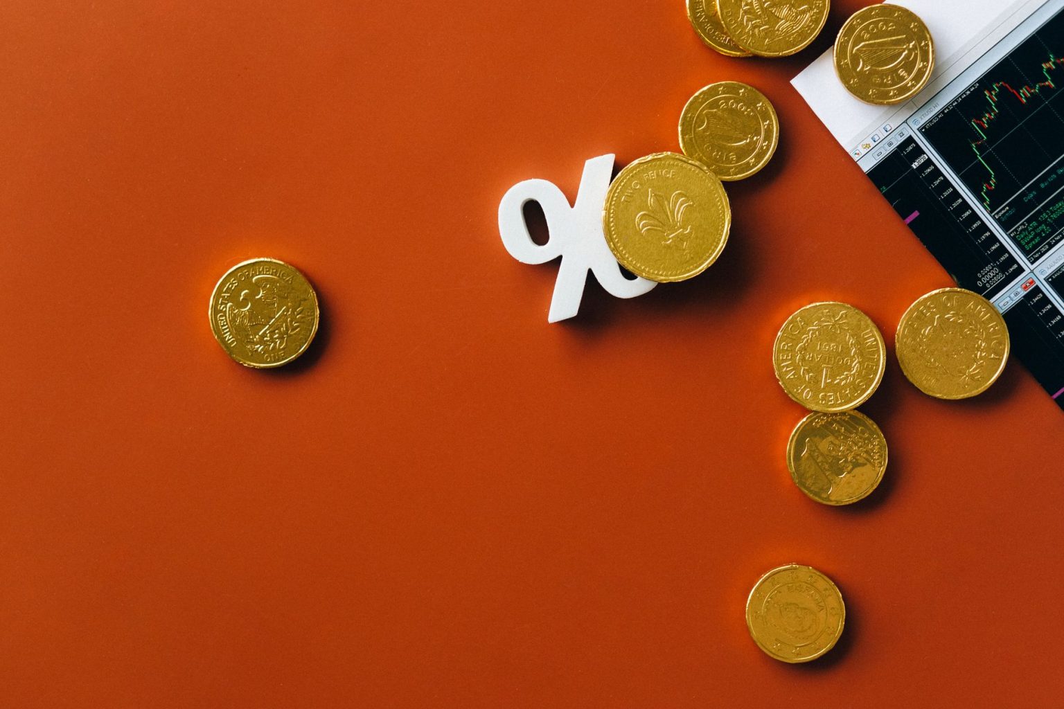 Moedas douradas sobre um gráfico de mercado, ao lado de um símbolo de porcentagem, em fundo laranja.