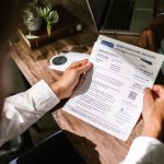 Homem consulta um documento financeiro sobre uma mesa de madeira, em ambiente iluminado por luz natural.