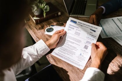 Homem consulta um documento financeiro sobre uma mesa de madeira, em ambiente iluminado por luz natural.
