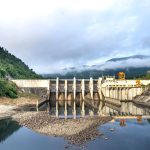 Usina hidrelétrica junto a um rio, com montanhas verdes ao fundo.