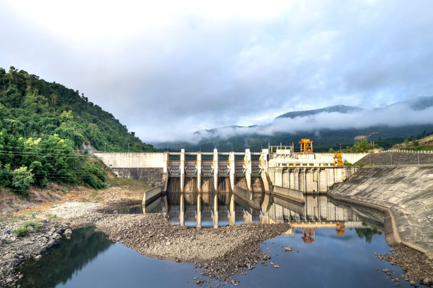 Usina hidrelétrica junto a um rio, com montanhas verdes ao fundo.